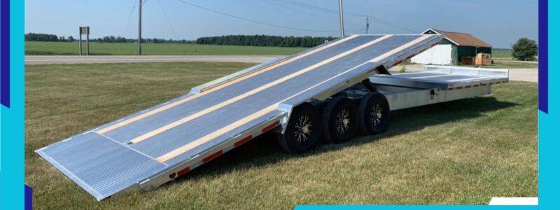 An aluminum tilt trailer parked on a grassy field, with its bed tilted downward, showcasing its loading functionality for heavy equipment.