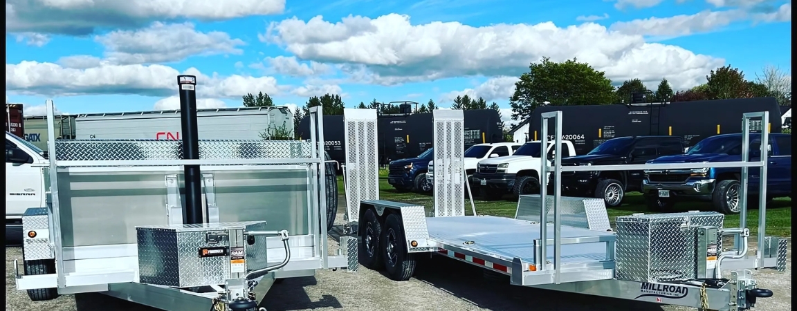 A selection of Millroad trailers, including a dump trailer and utility trailer, displayed outdoors with a backdrop of parked vehicles and blue skies.